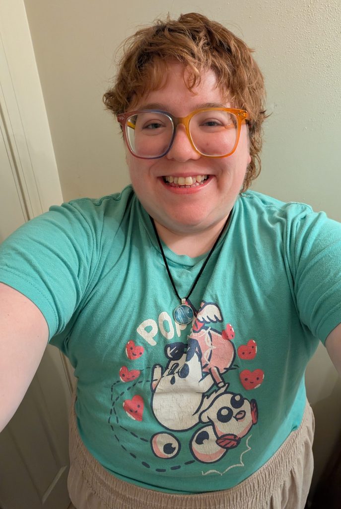 A nonbinary person with a reddish mullet wearing a mint green tee and rainbow-framed glasses.