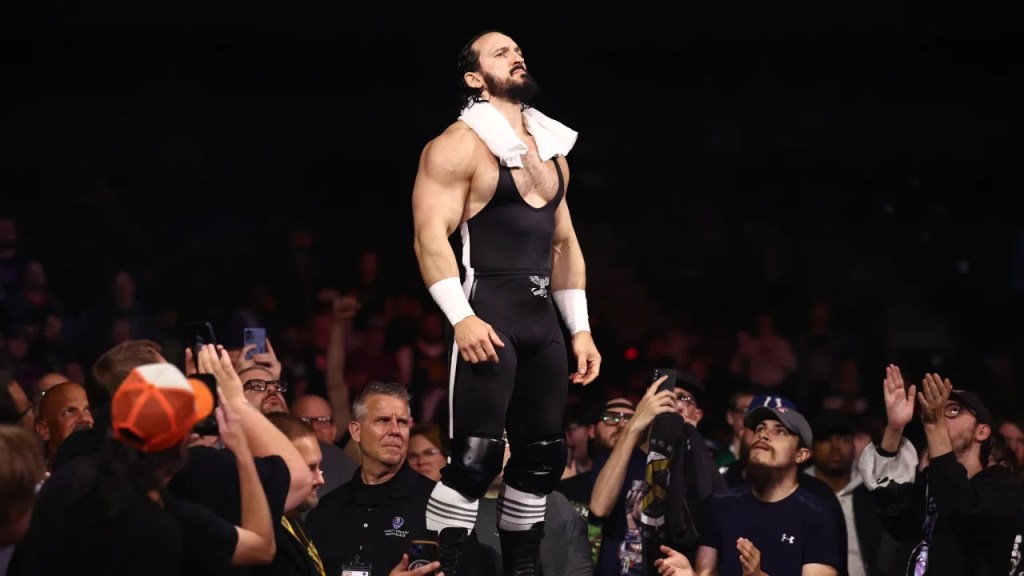 A wrestler in his gear, which includes a unitard, shorts, and knee pads, stands on the barricade surrounding the ring with a white towel around his neck. His chin is tilted up, and a crowd of admirers look up to him, some clapping, some taking photos and video with their phones.